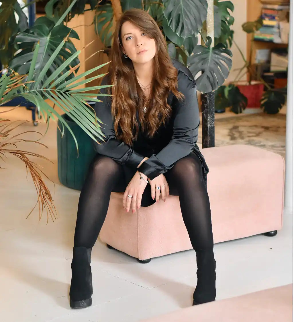 Une femme aux longs cheveux bruns, portant un haut en satin noir, des collants noirs et des bottines noires, assise sur un pouf rose clair.