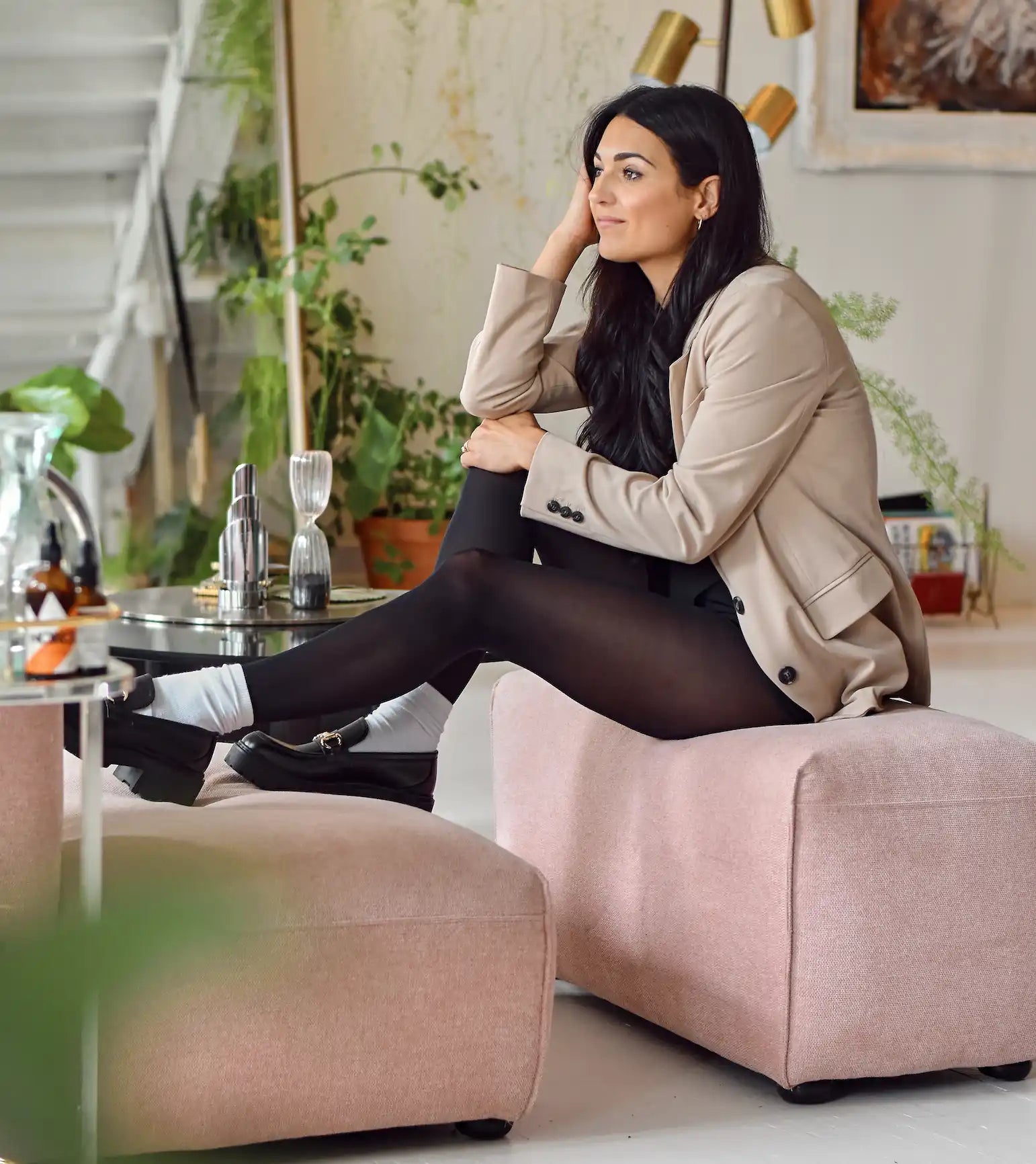 Une femme portant un blazer beige, des collants noirs, des chaussettes blanches et des chaussures noires, assise sur un banc rembourré rose.
